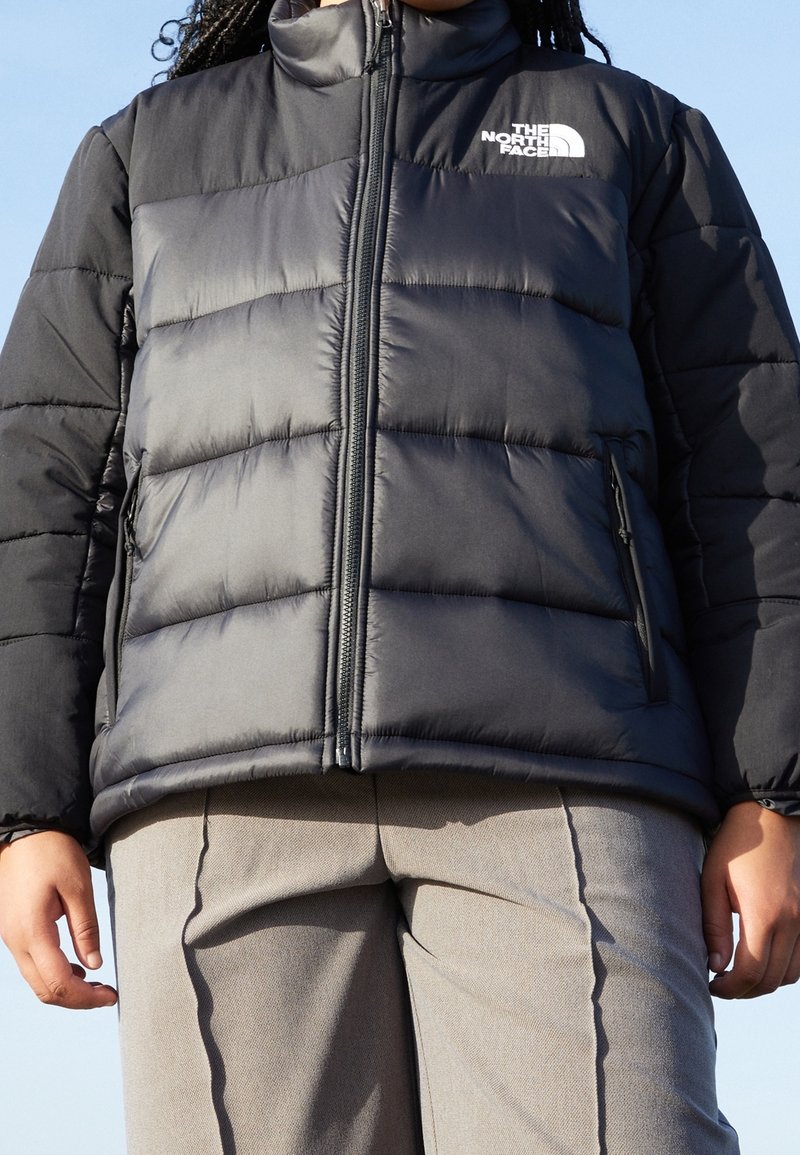 Doudoune noire avec col haut, design matelassé et logo blanc en haut à gauche. Poches zippées de chaque côté.