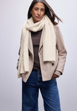 Beige wollen jas met een op maat gemaakt ontwerp en zwarte knopen, gecombineerd met een zachte, crèmekleurige sjaal. Het model draagt een donkere trui en blauwe spijkerbroek.