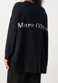 Czarna bluza z długimi rękawami, z dużym białym napisem na plecach, który brzmi "Marc O'Polo". Luźny krój, gładka tekstura.