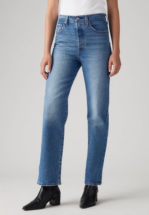 Personne portant un jean bleu droit taille haute, un haut blanc et des bottines noires, debout devant un fond gris clair uni.