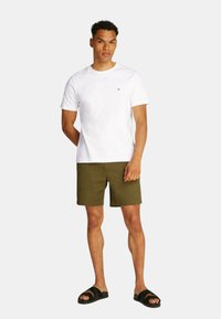 T-shirt bianco a maniche corte con un piccolo logo sul lato sinistro del petto, abbinato a pantaloni corti verde oliva e sandali neri con doppia fascia.