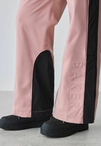 Rožinės ir juodos kelnės su blizgiu paviršiumi, turinčios šoninį išpjovimą ir reljefinį tekstą „BAKER“ šone, derinamos su juodomis platforminėmis bateliais.