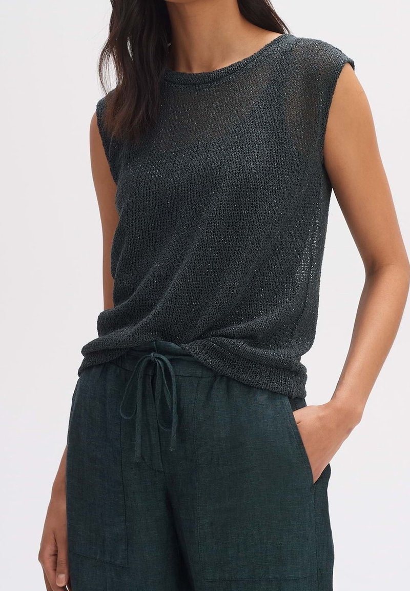 Femme portant un haut noir sans manches en maille et un pantalon foncé à cordon, une main dans la poche, sur un fond uni.
