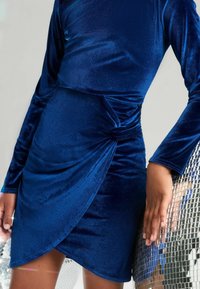 Mujer vestida con un vestido envolvente de terciopelo azul de manga larga, de pie al lado y tocando una gran bola de discoteca plateada.