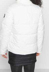 Vit vadderad jacka med hög krage, quiltad design och sömmar i sektioner. Har en logotypmärke på överarmen och en slät textur.