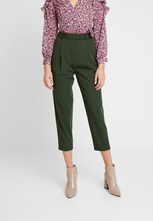 Vrouw die hooggetailleerde donkergroene cropped broek draagt, een roze blouse met bloemenprint en lange mouwen, en beige enkellaarsjes met blokhakken.