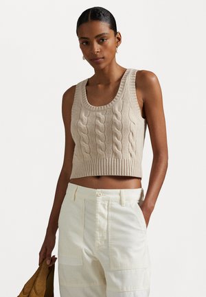 Top corto de punto beige con patrón de trenzado y dobladillo acanalado, combinado con pantalones crema de cintura alta. El modelo sostiene un bolso marrón.