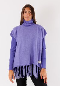 Poncho viola con collo a dolce e orlo frangiato, texture morbida, design a maniche corte e dettaglio con etichetta sul lato. Indossato sopra una maglietta a maniche lunghe.