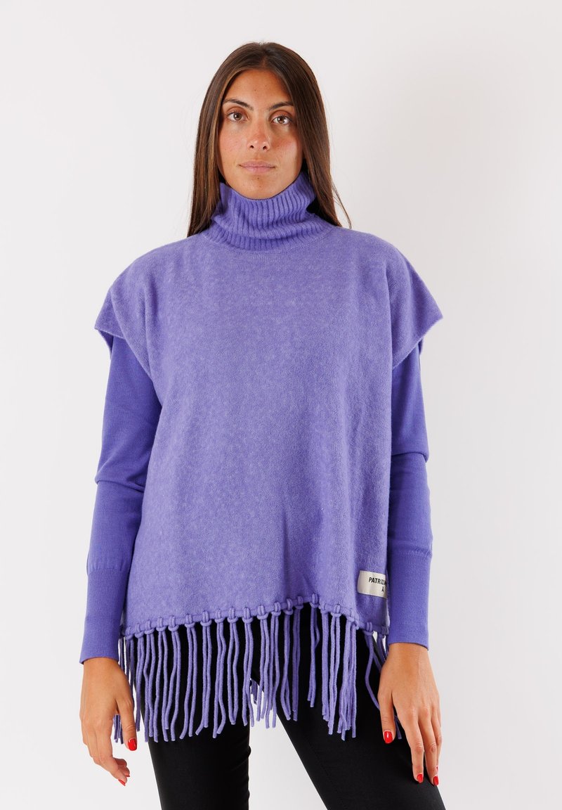 Poncho viola con collo a dolce e orlo frangiato, texture morbida, design a maniche corte e dettaglio con etichetta sul lato. Indossato sopra una maglietta a maniche lunghe.