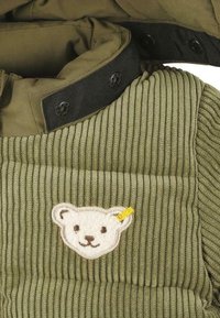 Gilet en tissu texturé vert avec des côtes verticales, comportant un patch représentant une tête d'ours avec un accent jaune. Détail de col noir avec des boutons.