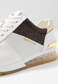 Sneaker bianco e marrone con un design perforato, dettagli in oro e suola trasparente. Il logo "MICHAEL KORS" è visibile sul lato.