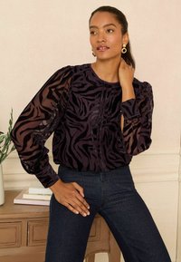 Blusa viola scura con maniche trasparenti, caratterizzata da un motivo a strisce zebra nere e dettagli con bottoni. Abbinata a jeans scuri.