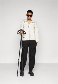 Witte textuurjack met capuchon en ritsluiting, gecombineerd met zwarte broek. Model houdt een wandelstok vast en draagt zwarte zonnebril.