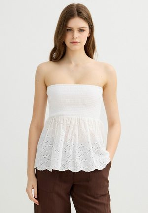 Joven con cabello largo y castaño viste un top blanco sin tirantes con bordados y pantalones marrones, de pie contra un fondo claro y liso.