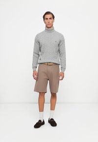Junger Mann steht und trägt einen grauen Rollkragenpullover aus Kabelstrick, einen beigen Gürtel, braune Shorts, weiße Socken und dunkelbraune Slipper vor weißem Hintergrund.