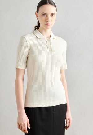 Ribbed, cream short-sleeve polo shirt with three button placket, fitted silhouette, and a soft texture. Worn with a black garment below.