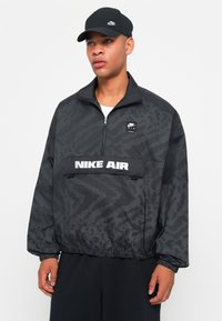 Svart glidelåsjakke med teksturert mønster, logo-patcher og bred midje. Kombinert med en matchende svart caps.