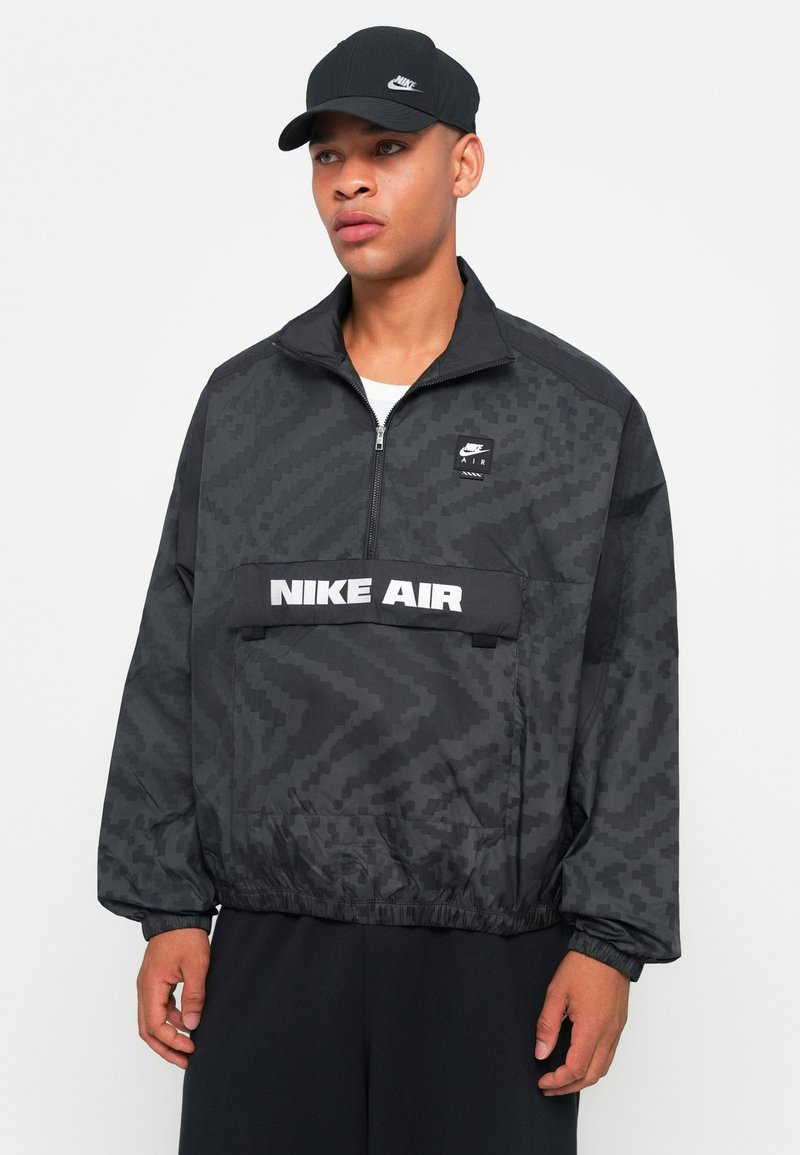 Chaqueta negra con cremallera que presenta un patrón texturizado, parches con logo y una amplia cinturilla. Combinada con una gorra negra a juego.