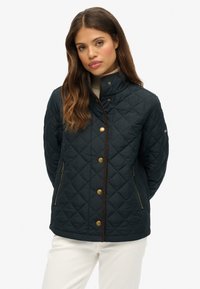 Veste matelassée navy avec quincaillerie dorée, col montant et poches latérales en diagonale ; elle présente une bordure marron le long de l'ouverture et des manches.