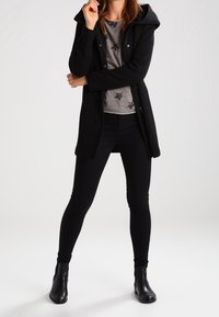 Abrigo negro con capucha y botones frontales, combinado con una camiseta gris con patrones de estrellas y jeans ajustados negros. Textura suave, corte moderno.