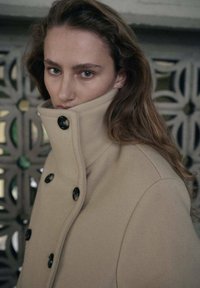 Beżowy wełniany płaszcz z dużymi czarnymi guzikami i wysokim kołnierzem. Gładka faktura, dopasowany fason i strukturalny krój. Modelka patrzy prosto w obiektyw.