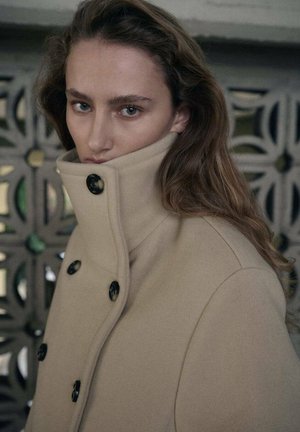 Beige wollen jas met grote zwarte knopen en hoge kraag. Gladde textuur, getailleerde pasvorm en gestructureerd ontwerp. Model kijkt recht in de camera.