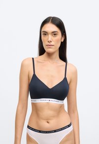 Czarny bezprzewodowy biustonosz z dekoltem w kształcie litery V i cienkimi ramiączkami. Posiada białą taśmę z logo "TOMMY HILFIGER". Gładka tekstura.