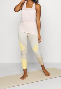 Světle růžový top na ramínka spárovaný s texturovanými béžovými legínami s žlutými a šedými úhlovými vzory, stojící na jógové podložce.