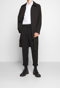 Černý dlouhý kabát přes bílou košili, doplněný černými kalhotami a černými botami. Vyznačuje se štíhlým designem a minimalistickým stylem.