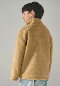 Béžová fleecová bunda s vysokým límcem, texturovaným povrchem a uvolněným střihem. Dlouhé rukávy a rovný lem dokončují design.