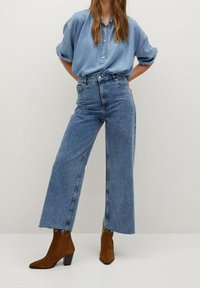 Chemise bleu clair à boutons avec manches retroussées, associée à un jean bleu taille haute à jambes larges. Bottines marron à talons avec bouts pointus.