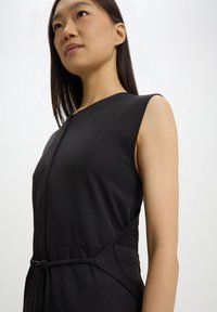 Abito nero senza maniche con corpino aderente, scollatura a V, tasche laterali e una cintura annodata in vita, realizzato in tessuto liscio e elasticizzato.
