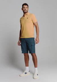 Geel polo shirt met een zwart logo, gecombineerd met teal cargoshorts met zijzakken en witte sneakers met blauwe accenten.