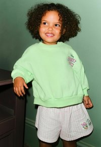 Világoszöld pulóver bordázott mandzsettákkal, rózsaszín grafikával. Fehér rövidnadrággal párosítva, amely rózsaszín rácsmintás és karakteres nyomattal rendelkezik.