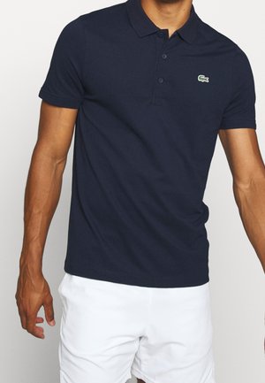 Mann, der ein dunkel marineblaues Polohemd mit kleinem grünem Krokodil-Logo auf der linken Brust und weißen Shorts trägt.