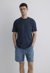 Marineblaue Baumwoll-T-Shirt mit Rundhalsausschnitt und kurzen Ärmeln, mit einem kleinen roten Emblem auf der linken Brust. Tragen mit Jeansshorts.