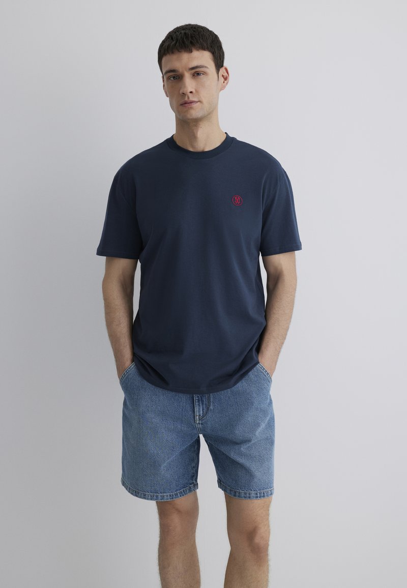 Marineblaue Baumwoll-T-Shirt mit Rundhalsausschnitt und kurzen Ärmeln, mit einem kleinen roten Emblem auf der linken Brust. Tragen mit Jeansshorts.