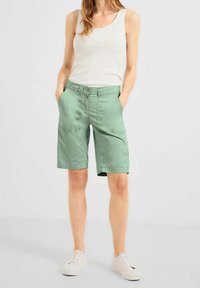 Shorts en coton verts à coupe décontractée, avec deux poches avant, une double fermeture à boutons et un ourlet replié. Portés avec un débardeur blanc et des baskets.