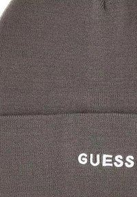 Grijze gebreide beanie met een geribbelde textuur. Heeft een omgeklapte rand en een wit "GUESS" logo aan de voorkant. Zacht, rekbaar materiaal.
