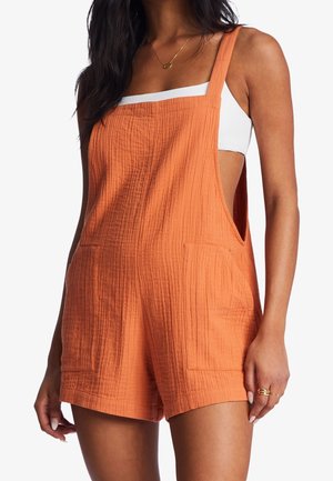 Frau trägt einen locker sitzenden, strukturierten orangen ärmellosen Overall mit Fronttaschen über einem weißen ärmellosen Oberteil, zeigt dabei nackte Arme und Beine.