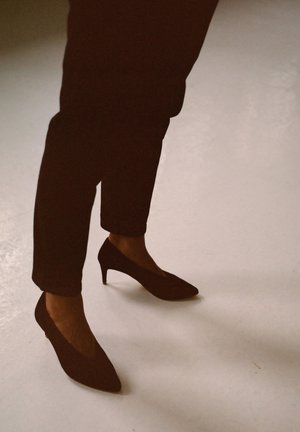Tacchi alti in suede neri con punte affusolate, posizionati su pantaloni scuri. I tacchi misurano circa 5 cm di altezza e presentano un design semplice.
