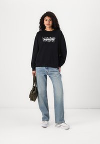 Felpa nera con logo "Levi's" in bianco, abbinata a jeans di denim azzurri chiari. Il modello tiene una piccola borsa verde oliva, in piedi contro uno sfondo neutro.