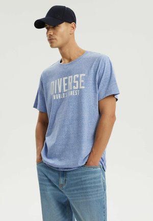 Młody mężczyzna w czarnej czapce, jasno niebieskiej koszulce z napisem "Diverse World's Finest" oraz niebieskich jeansach, dłonie w kieszeniach, spogląda na bok.