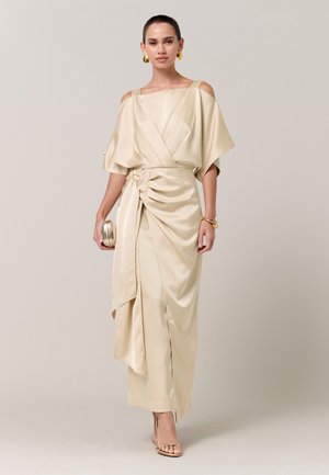 Vrouw draagt een elegante satijnen jurk in champagnekleur met cold-shoulder mouwen, houdt een bijpassende clutch vast en draagt gouden sieraden.