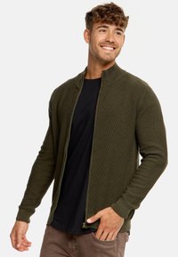 Cárdigan de punto texturizado en verde oliva con cremallera, que presenta un alto cuello y puños ajustados. Estilizado sobre una camiseta negra.