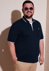 Námornícka polokošeľa s bielym golierom a lemovaním, krátkymi rukávmi a detailom loga. Nosená s svetlými béžovými nohavicami. Svetlá textúra a voľný strih.