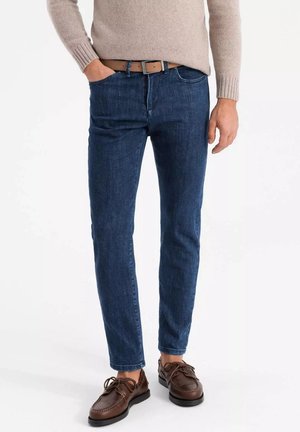 Donkerblauwe spijkerbroek met een slim fit, met vijf zakken en een riem. Gecombineerd met bruine leren schoenen en een beige gebreide trui.