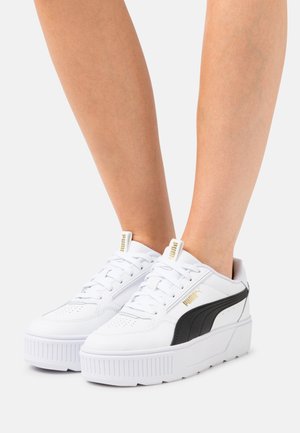 Zapatillas blancas con parte superior de cuero texturizado, panel de acento negro, gruesa suela de plataforma blanca y detalle del logo dorado en el costado.