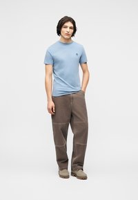 Νέος άνδρας στέκεται με το ένα χέρι στην τσέπη, φοράει ανοιχτό μπλε κοντομάνικο πουκάμισο, καφέ χαλαρό παντελόνι και μπεζ μπότες σε απλό φόντο.