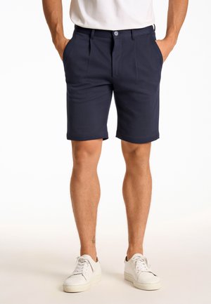 Mand iført marineblå skræddersyede shorts med frontplisser, hvide sneakers og en hvid t-shirt, hænderne i lommerne, stående på en lys baggrund.